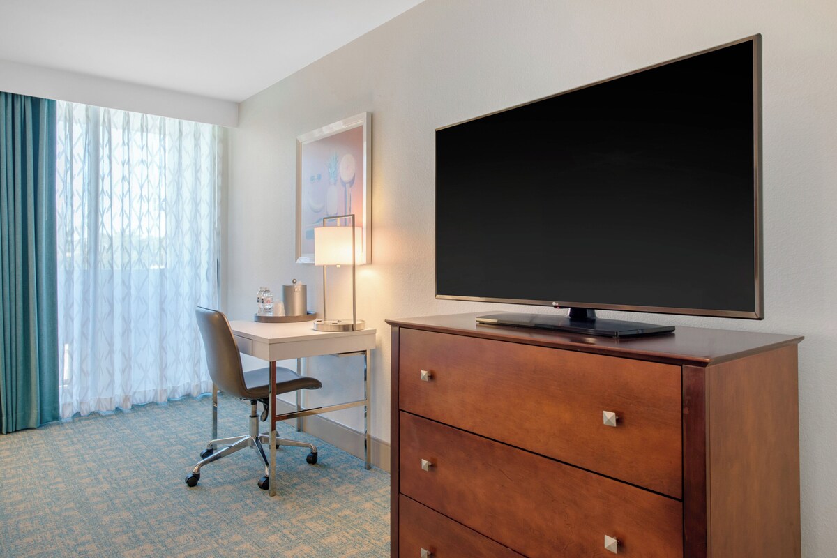 A hotel room with a desk, chair, TV, and a dresser.
