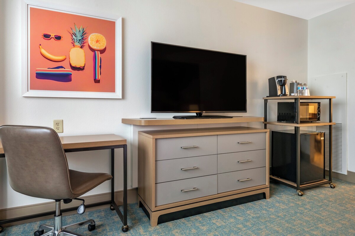 A hotel room with a desk, chair, television, minibar, and a framed picture on the wall.
