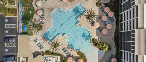 An aerial view of a hotel with a large swimming pool, sun loungers, and a parking area.
