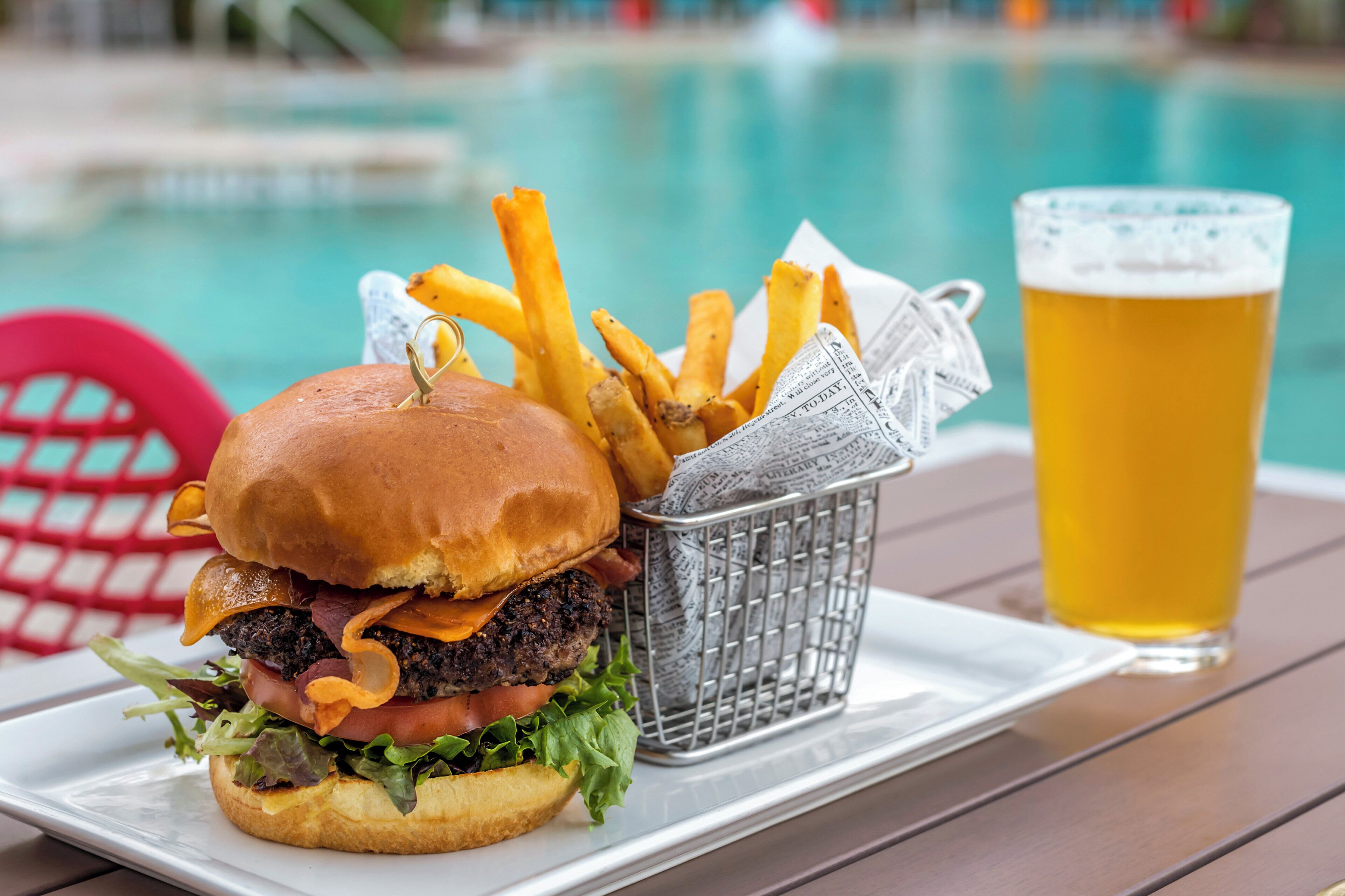 A burger, fries, and a glass of beer on a table by a pool.