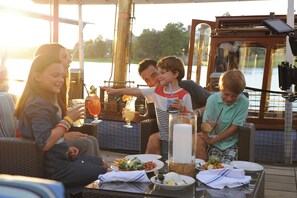 A group of people seated at an outdoor dining area with a view of a body of water, surrounded by wicker furniture and various dishes.