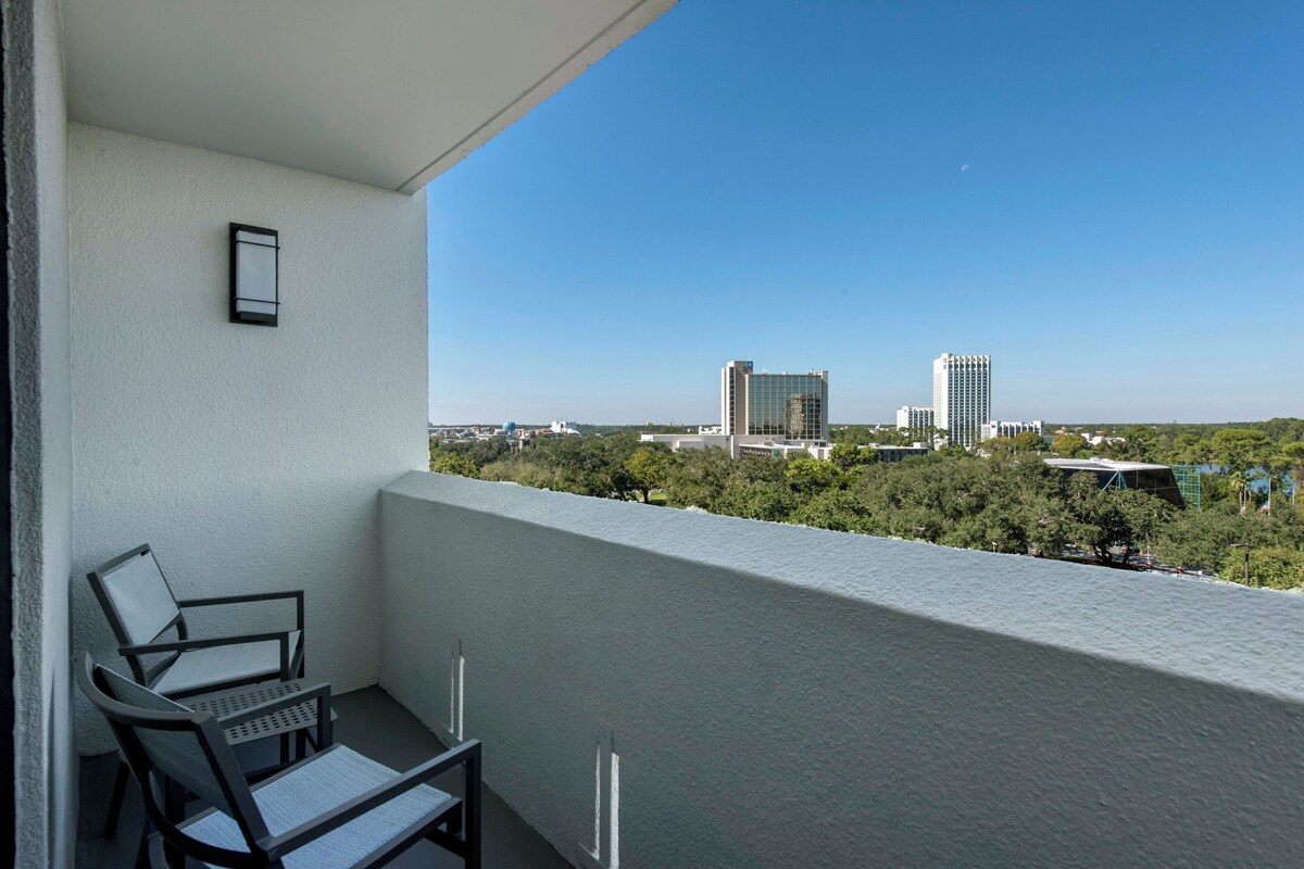 A balcony with two chairs, a small table, and a view of city buildings and greenery.