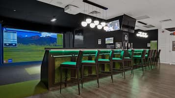 A golf simulation room with a large screen displaying a golf game, multiple screens, and a bar area with green chairs.