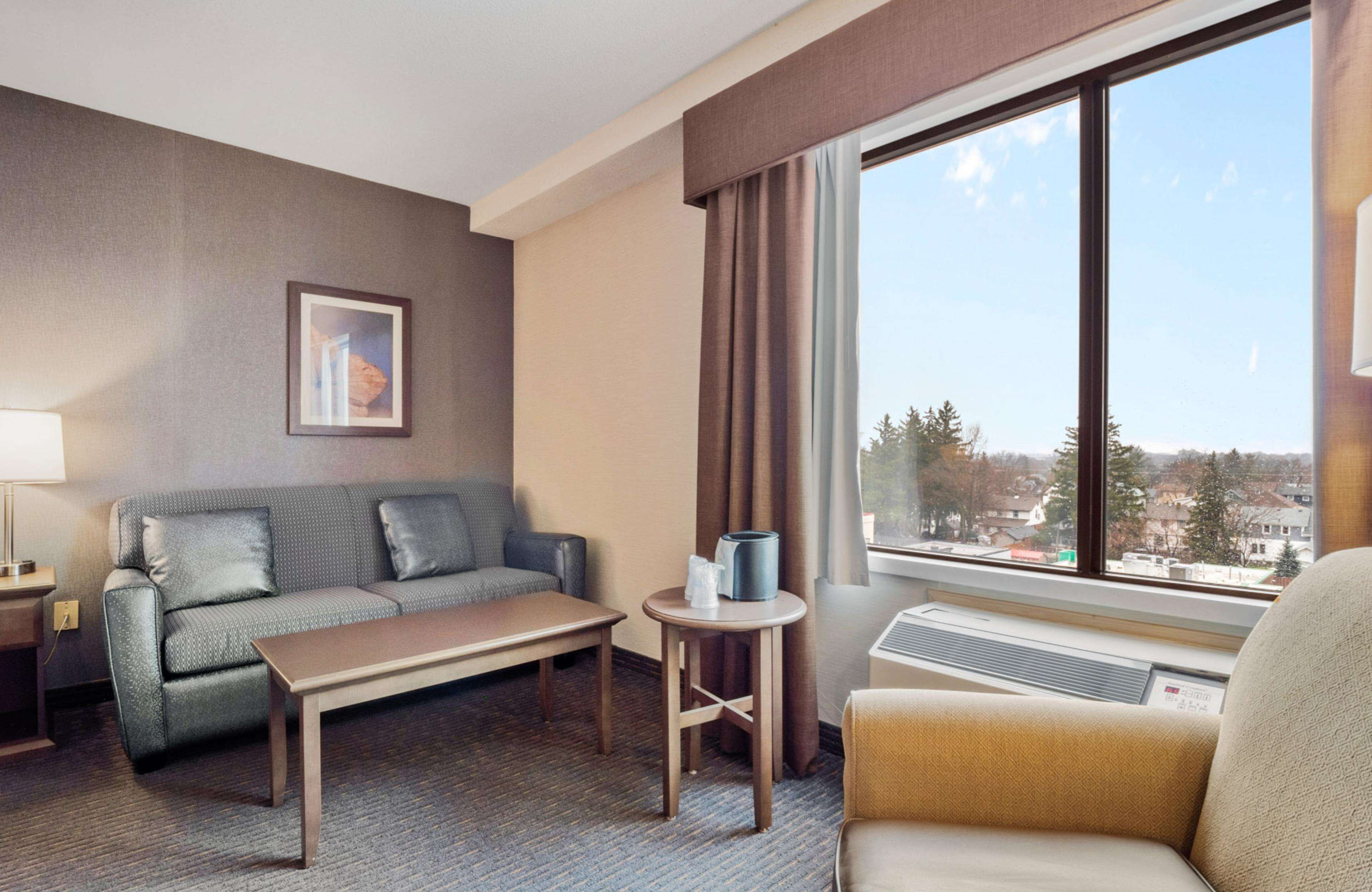 Hotellihuoneessa on harmaa sohva, puinen pöytä, pieni pyöreä pöytä, jossa on lamppu, ja tuoli.