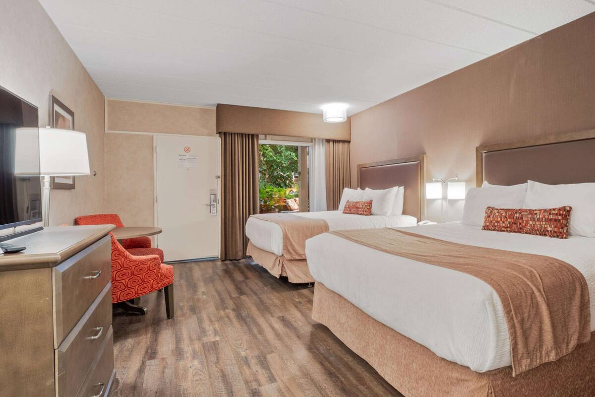 A hotel room with two beds, a desk with a lamp, a chair, and a view of greenery outside.