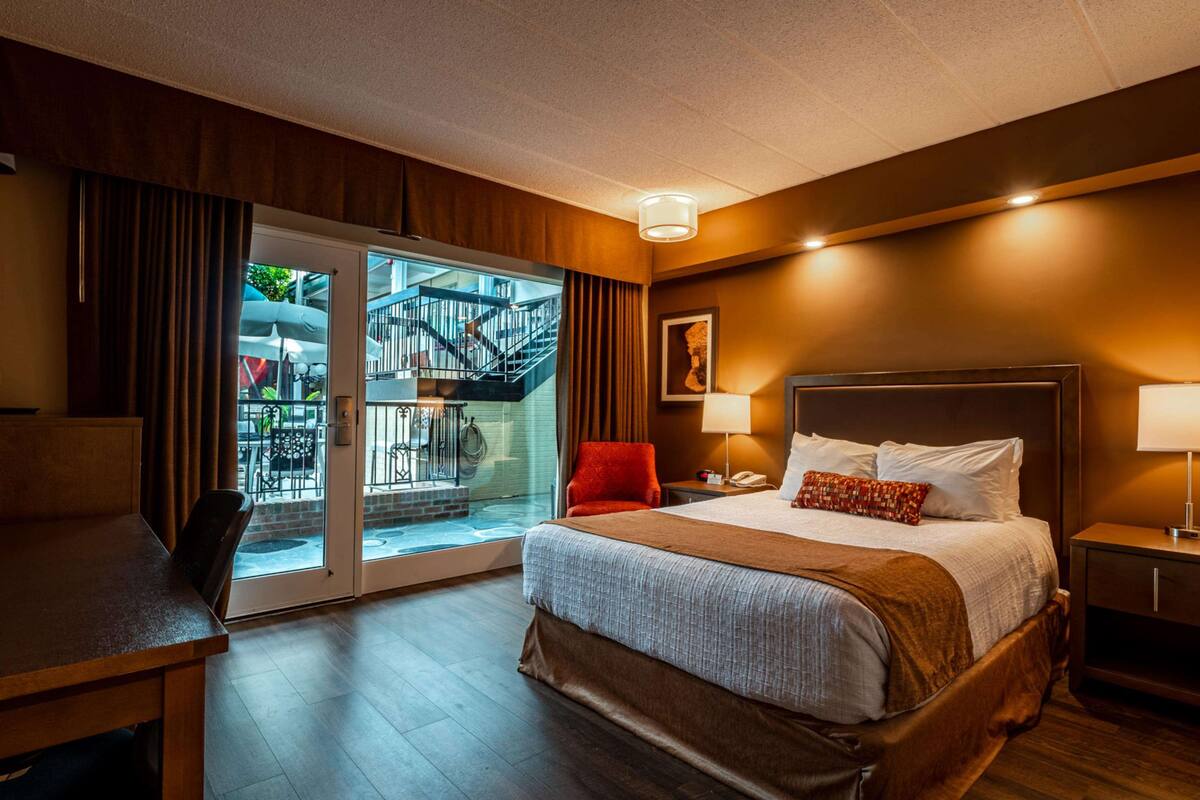A hotel room with a large bed, a desk, a chair, a nightstand, and a view of a balcony with a staircase.