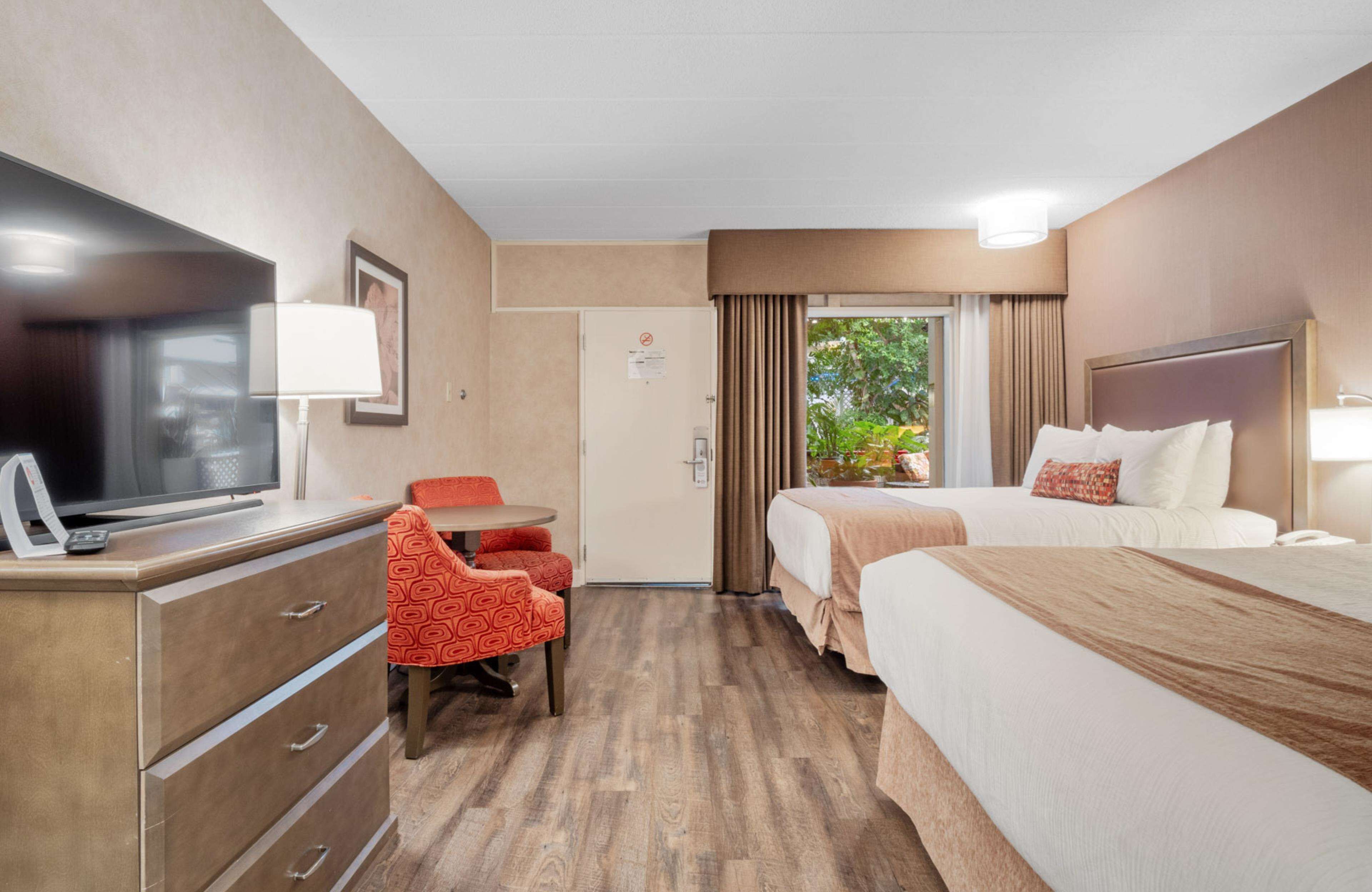 Habitación de hotel con televisor, escritorio con silla, dos camas y puerta que da a un balcón con vistas a zonas verdes.