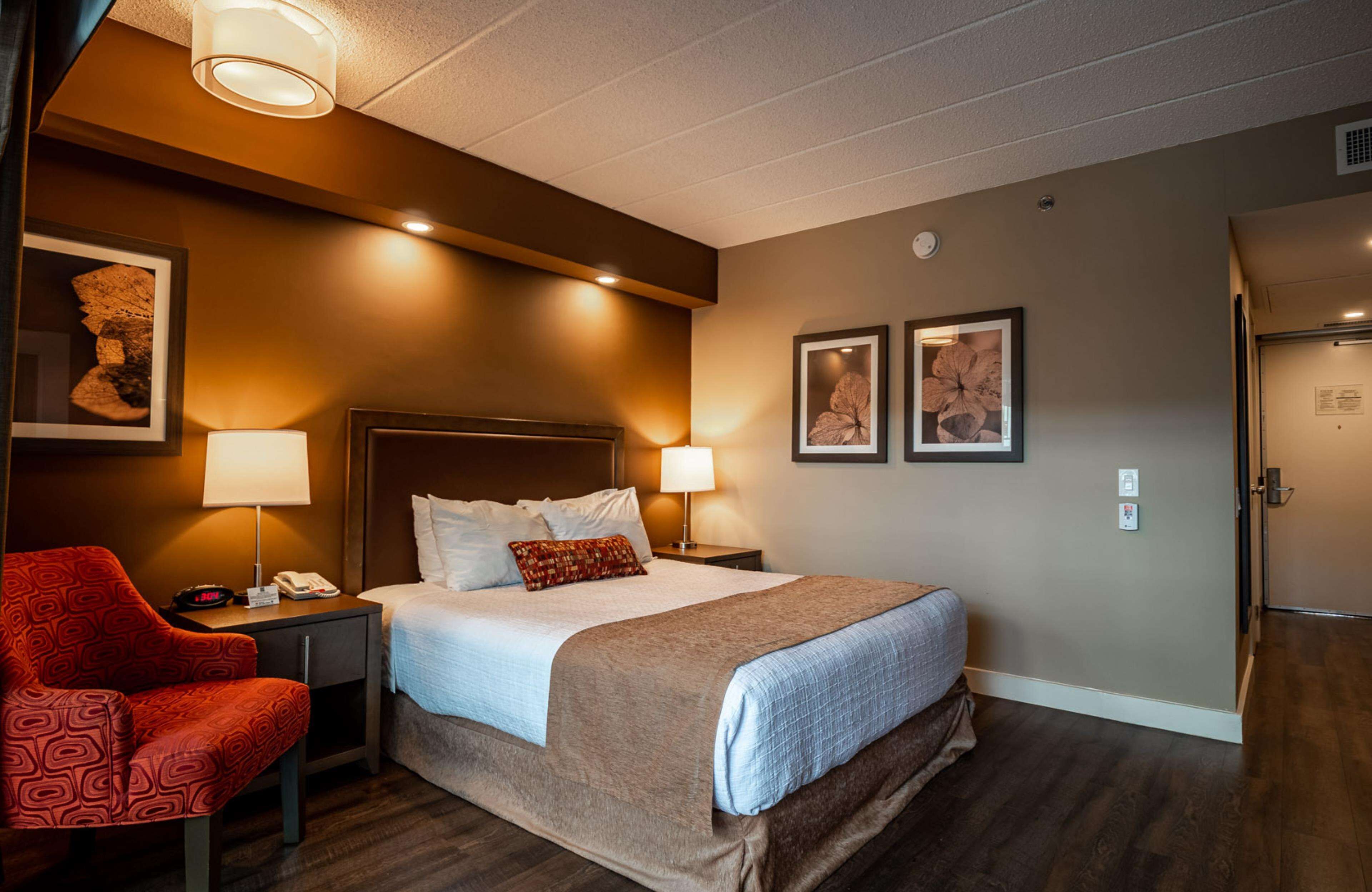 A hotel room with a bed, two bedside lamps, a red chair, and two framed pictures on the wall.