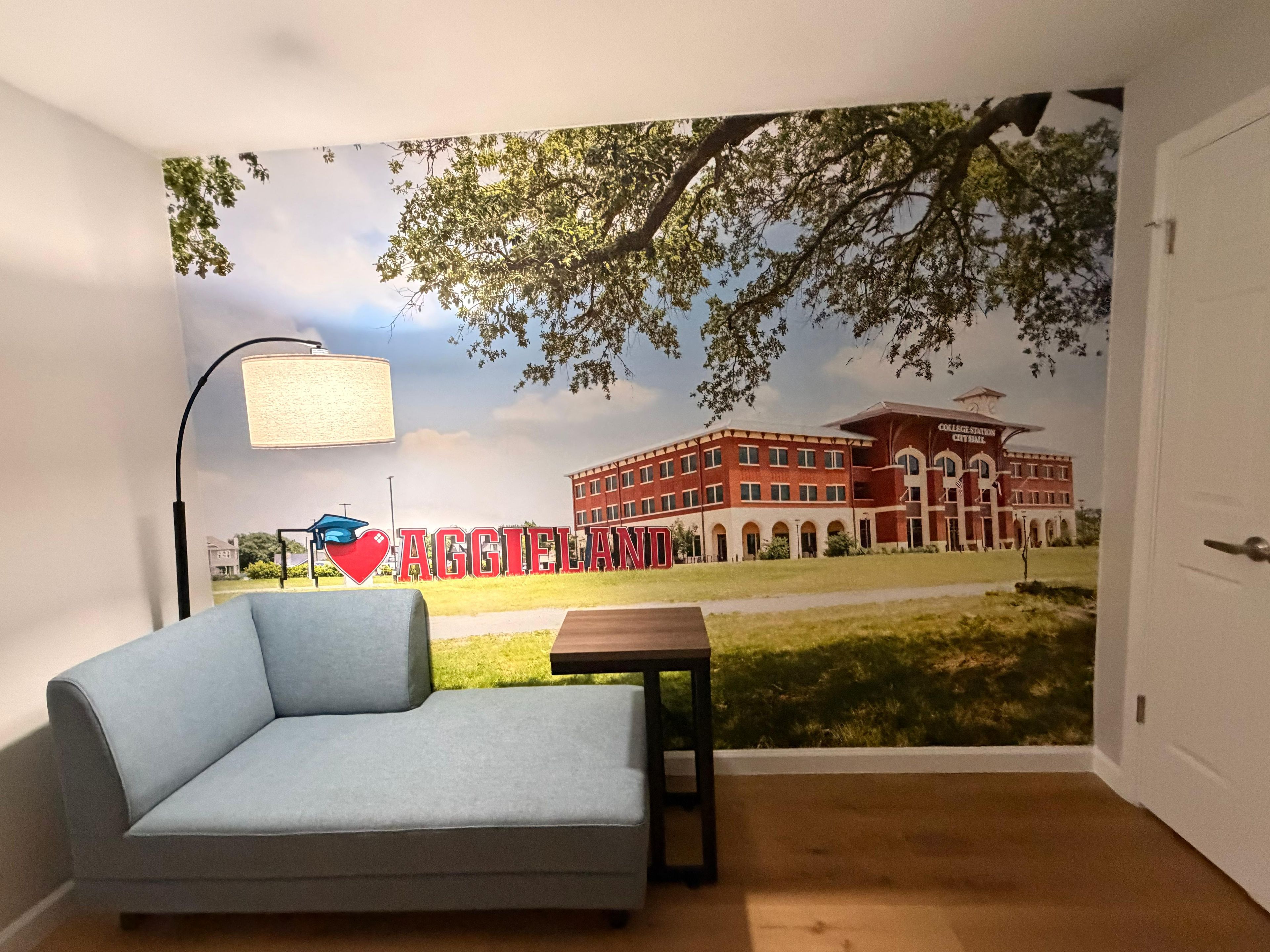 A room with a blue sofa, a small table, a lamp, and a mural of a building with the word 'AGGIELAND' on it.
