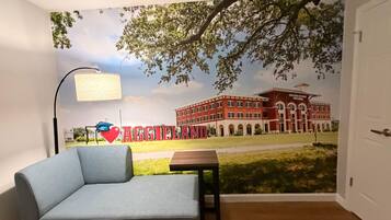 A room with a blue sofa, a small table, a lamp, and a mural of a building with the word 'AGGIELAND' on it.