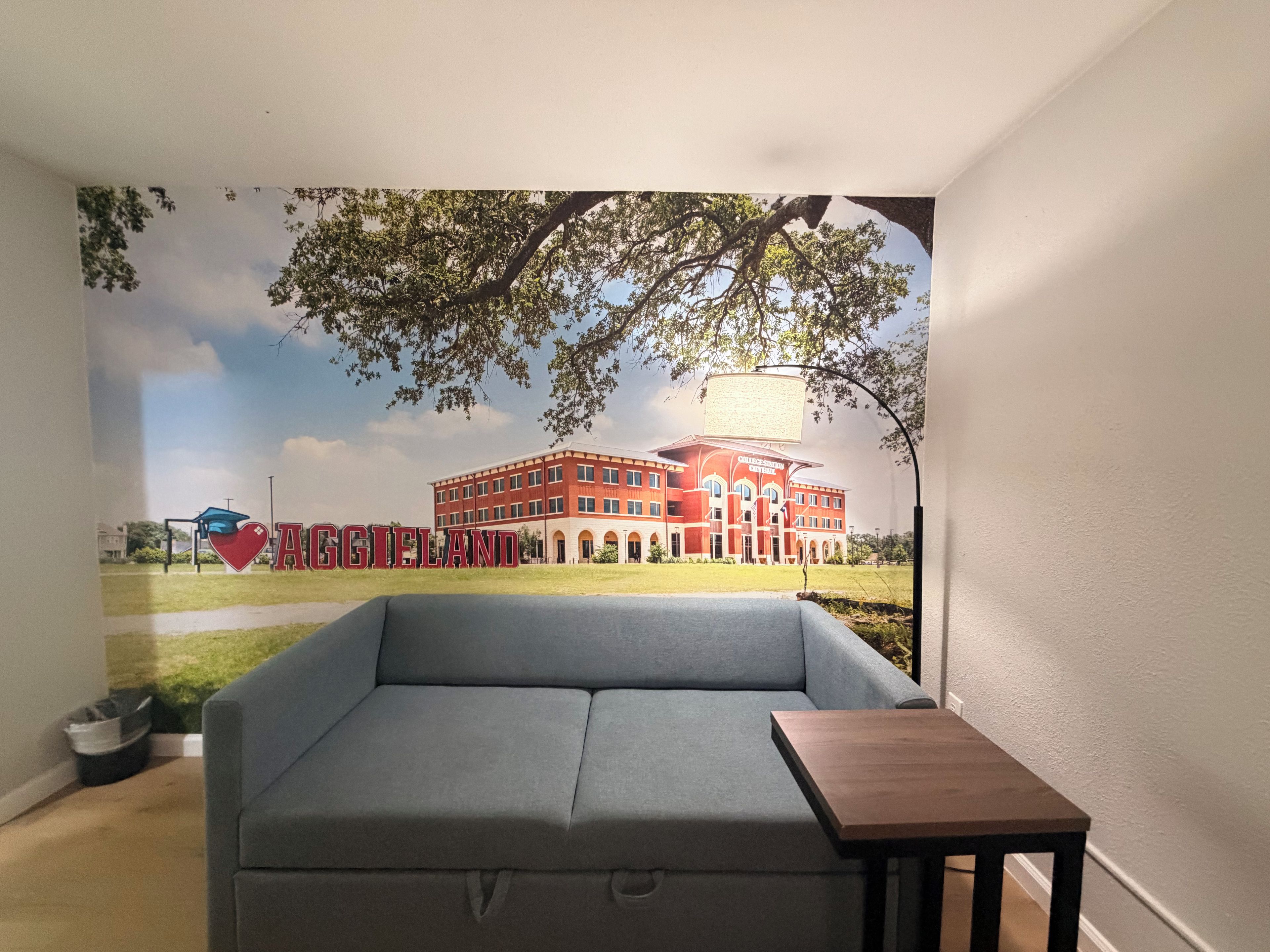 A room with a grey sofa, a small wooden table, and a wall mural of a building with a tree.