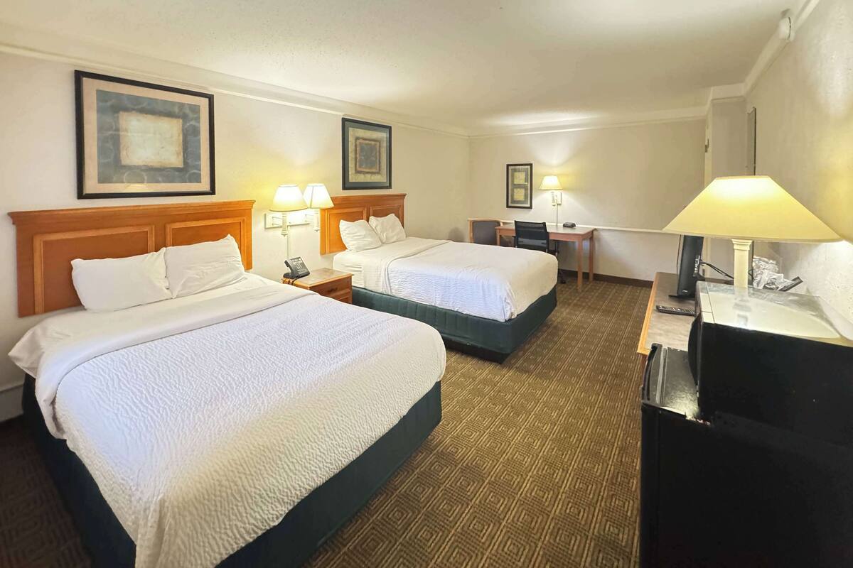 A hotel room with two beds, a desk, and a chair. There are lamps on the desk and bedside tables.