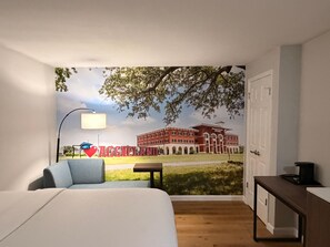 A bedroom with a large bed, a desk, a lamp, and a mural of a building with the word 'AGGIELAND' on it.