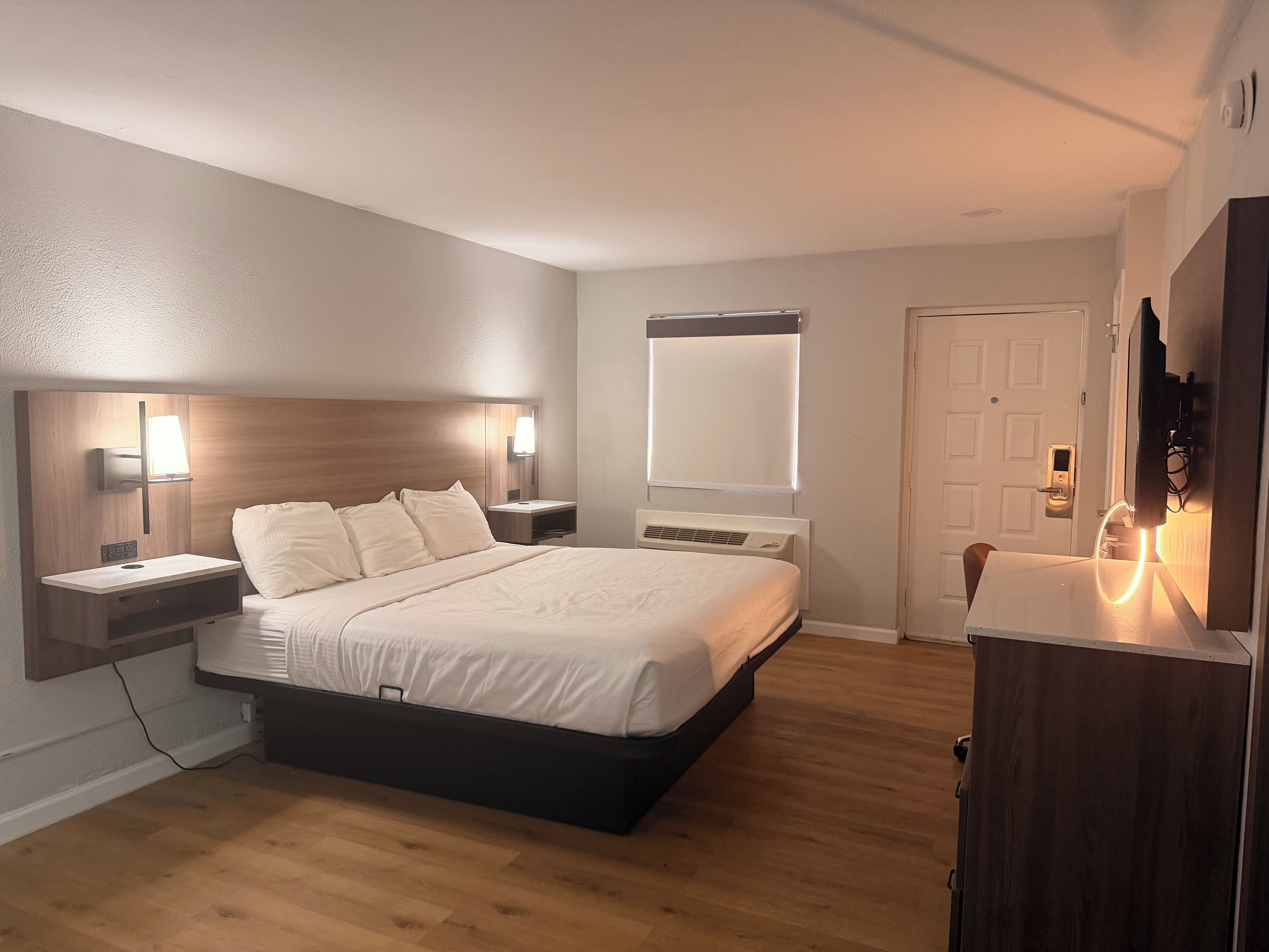 A hotel room with a bed, bedside tables, a nightstand, a mirror, a door, and a desk with a lamp.