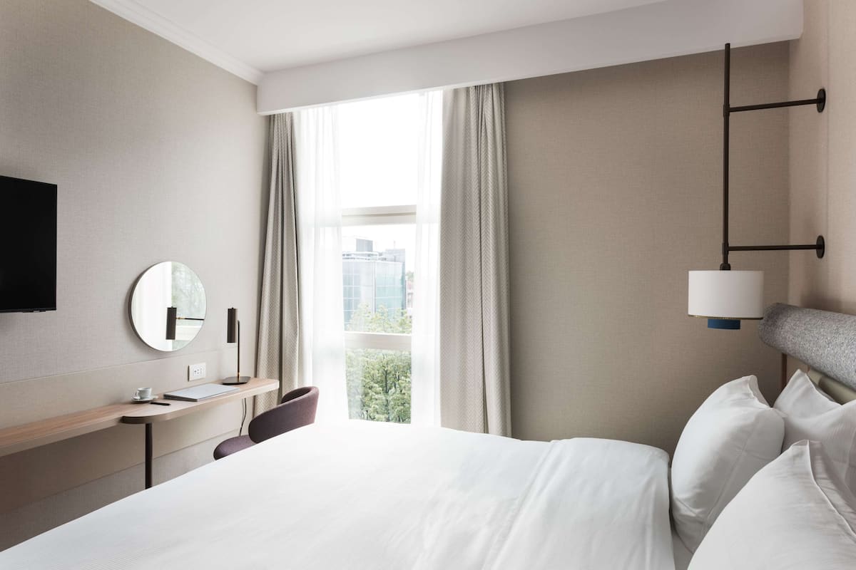 A hotel room with a bed, a desk, a chair, a round mirror, a flat-screen TV, and a window with curtains.