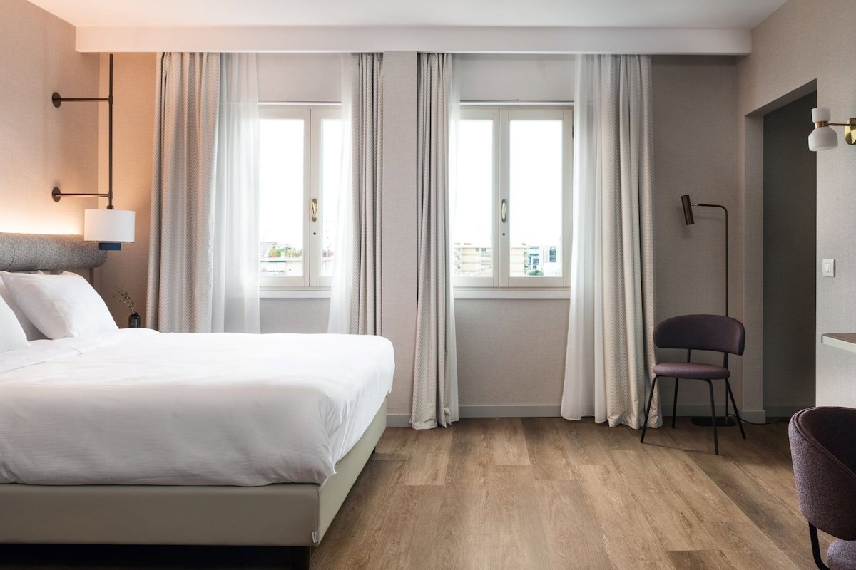 Habitación de hotel con cama, una silla, una lámpara de pie y vistas a la ciudad a través de la ventana.