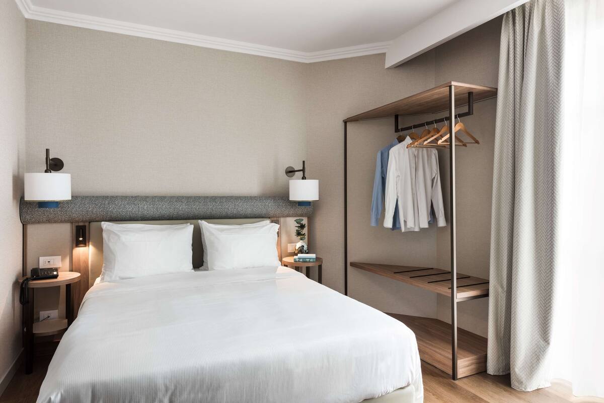 A hotel room with a large bed, bedside lamps, a small table with a phone, and a closet with hanging clothes.
