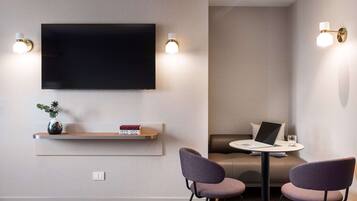 Una habitación de hotel moderna con una televisión de pantalla plana montada en la pared, una mesita con una computadora portátil y un vaso, dos sillas moradas, un sofá y una estantería con objetos decorativos.