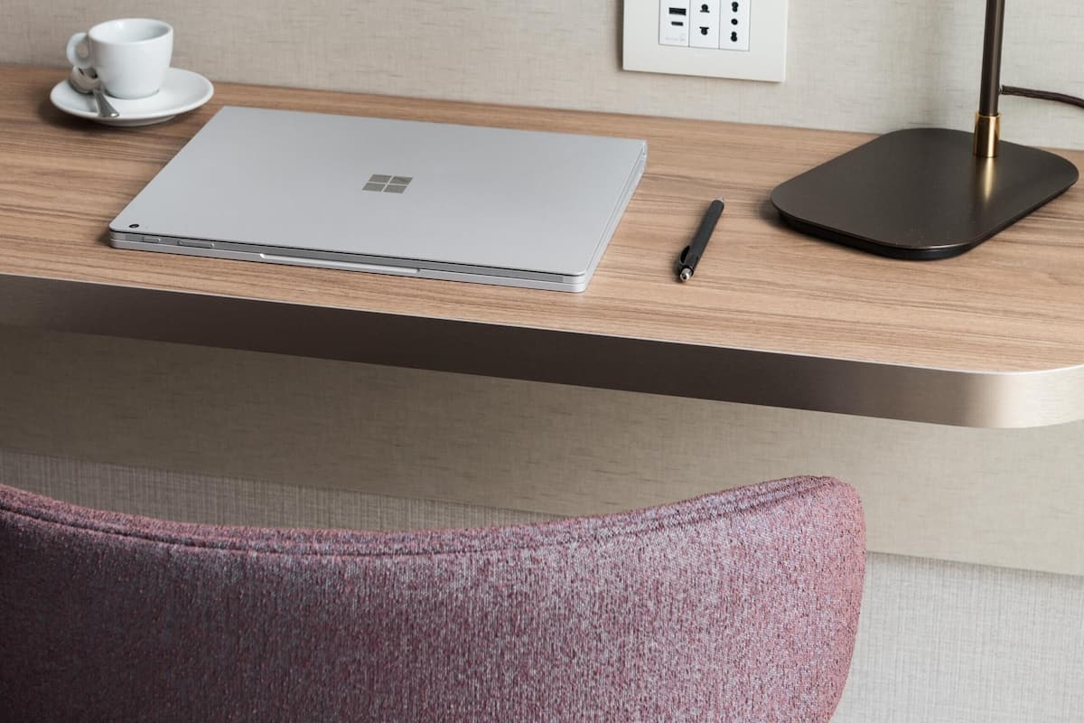 A neatly arranged hotel room with a round mirror, a laptop, a cup, and a pen on a wooden desk.