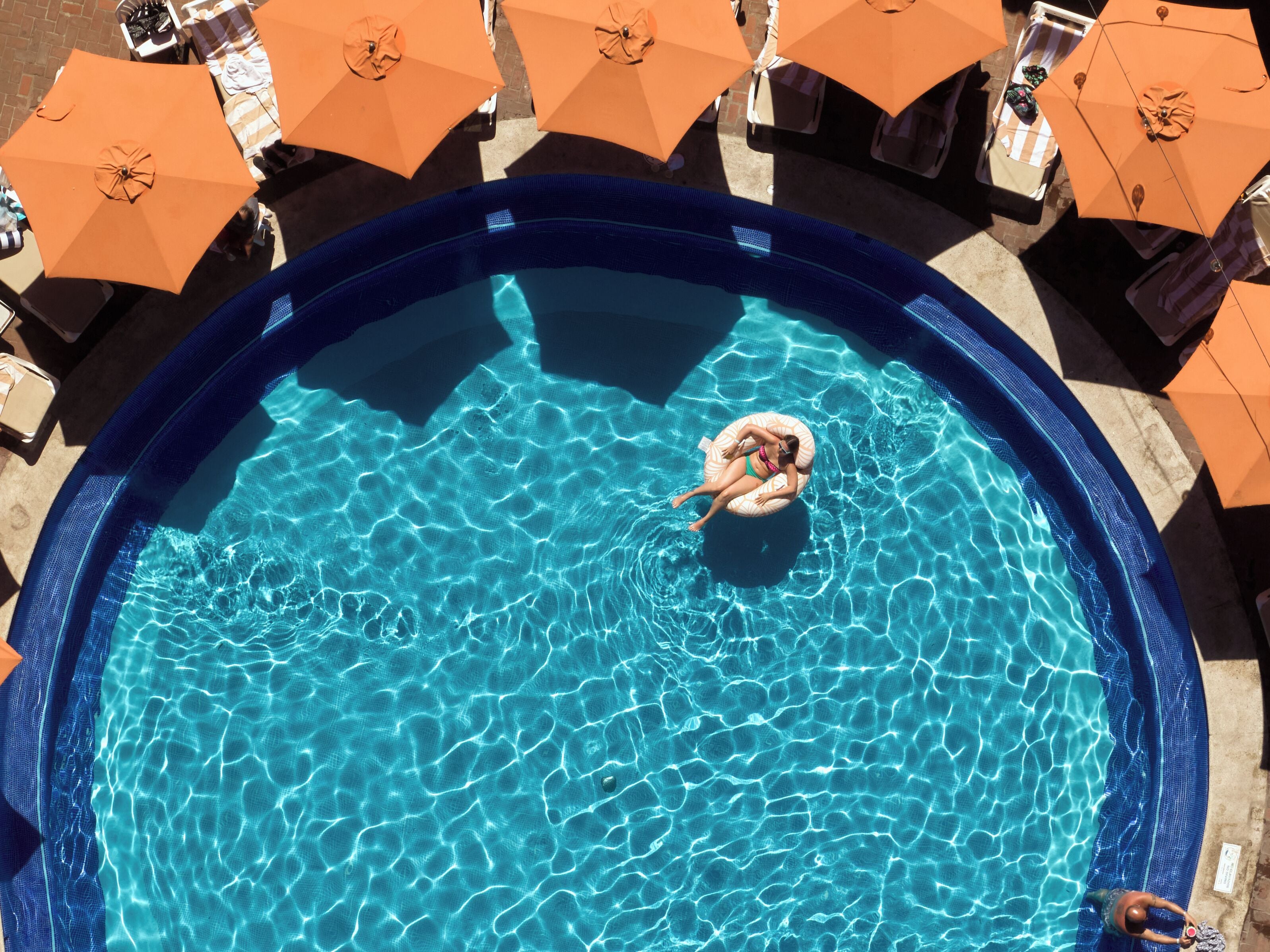 En swimmingpool med en person, der flyder på en luftmadras, omgivet af orange parasoller og liggestole.