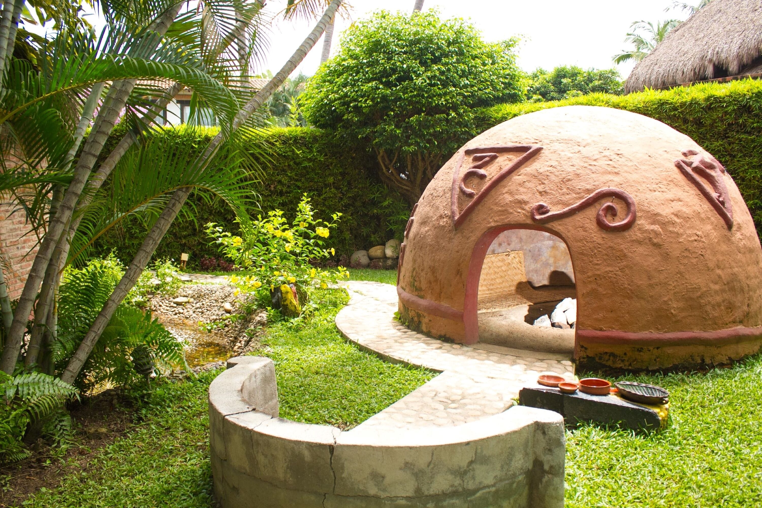 A traditional clay oven with a stone pathway leading to it, surrounded by lush greenery and palm trees.