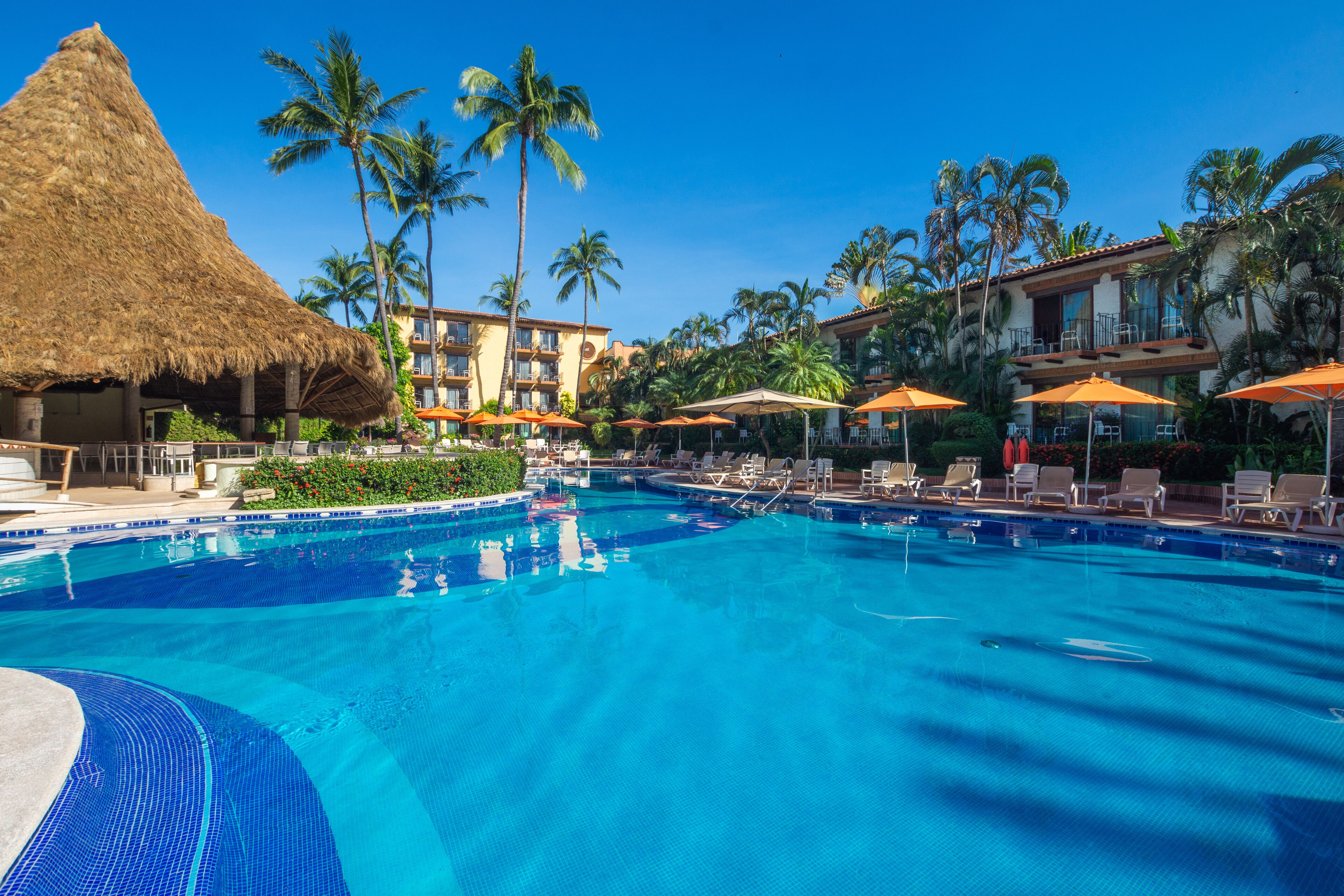 A swimming pool with lounge chairs and umbrellas, surrounded by palm trees and a building with balconies.