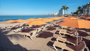 Una playa con filas de tumbonas y sombrillas naranjas, un cielo azul despejado y un paisaje urbano al fondo.