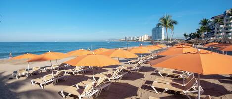 Una playa con filas de tumbonas y sombrillas naranjas, un cielo azul despejado y un paisaje urbano al fondo.