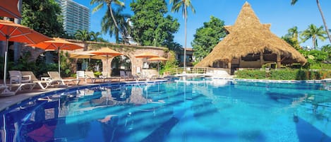 A swimming pool with lounge chairs and umbrellas, a thatched-roof structure, and palm trees.