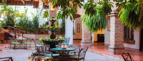 An indoor dining area with tables and chairs, surrounded by lush greenery and hanging plants.