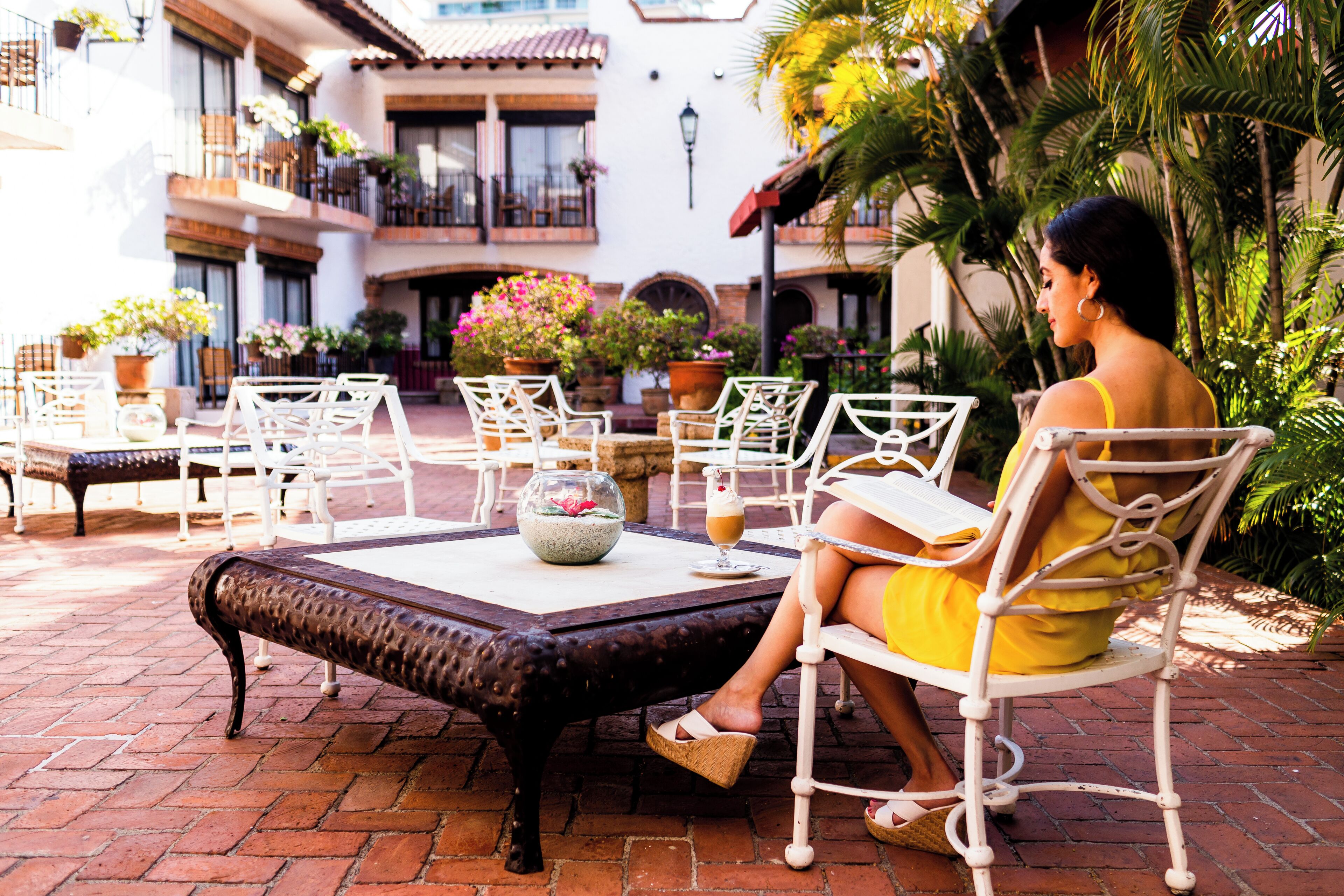 A woman seated at a table in a courtyard with potted plants and outdoor furniture.