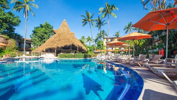 Una piscina con sillas de descanso y sombrillas, rodeada de palmeras y una estructura con techo de paja.