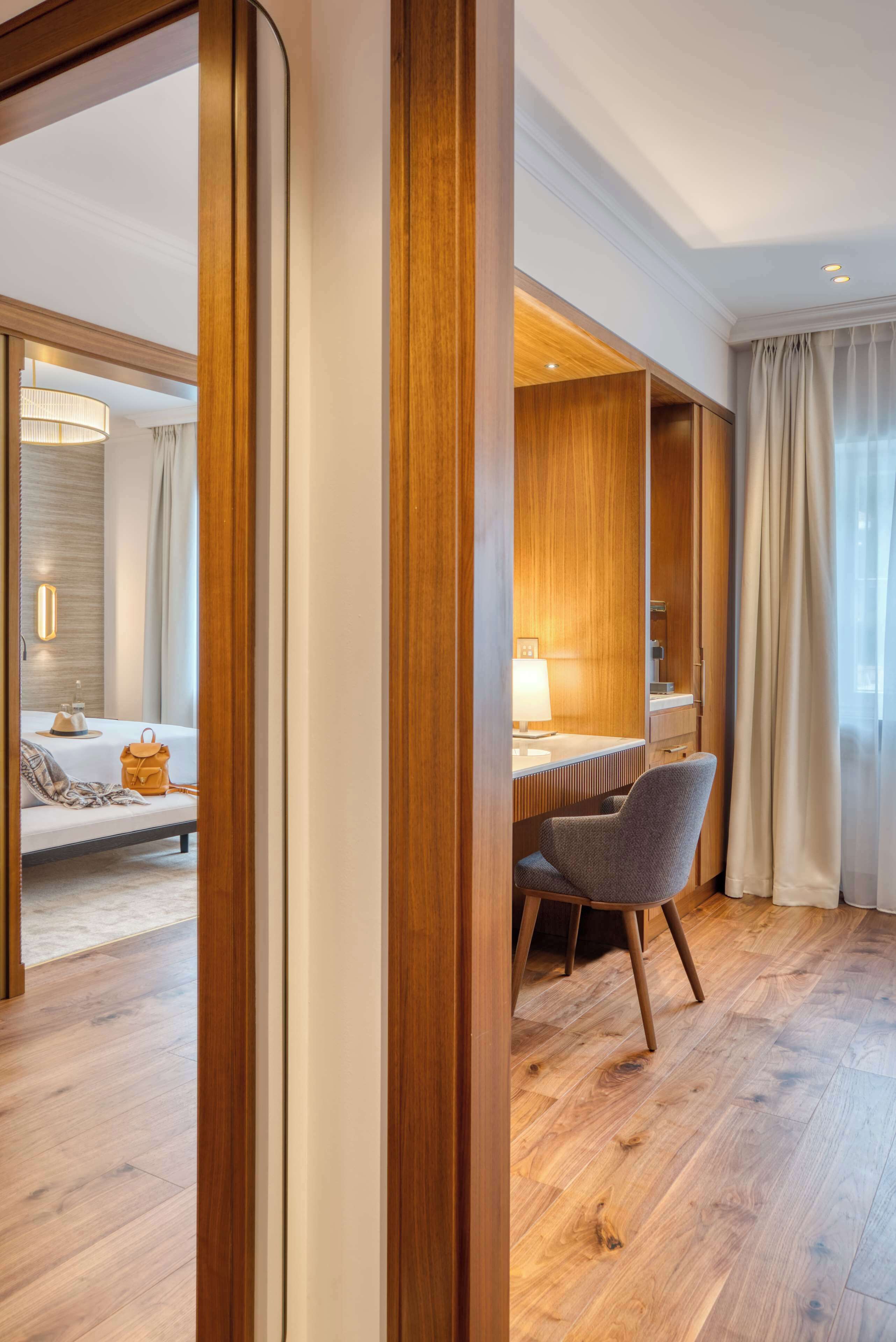 A hotel room with a wooden floor, a bed with a white headboard, a desk with a lamp, and a chair.
