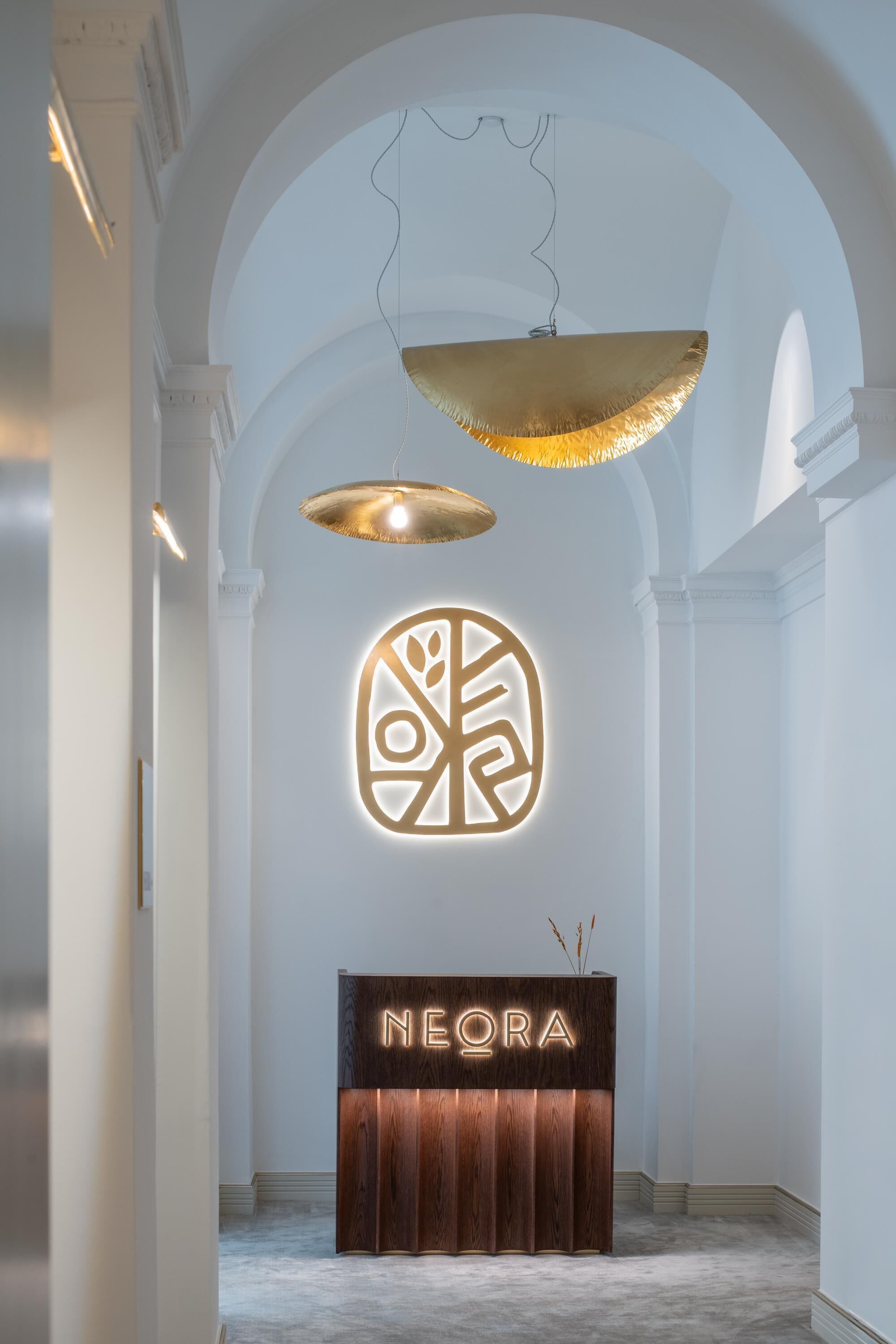 Un'area reception con una scrivania in legno con l'etichetta "NEORA", un logo illuminato a parete e due lampade a sospensione.