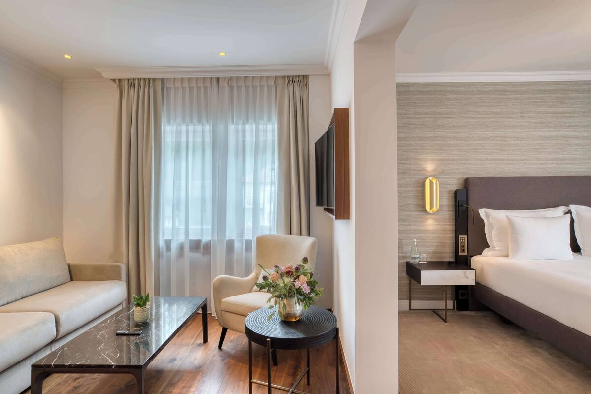 Una habitación de hotel moderna con sofá, sillón, una pequeña mesa redonda con un jarrón de flores, cama y televisor montado en la pared.