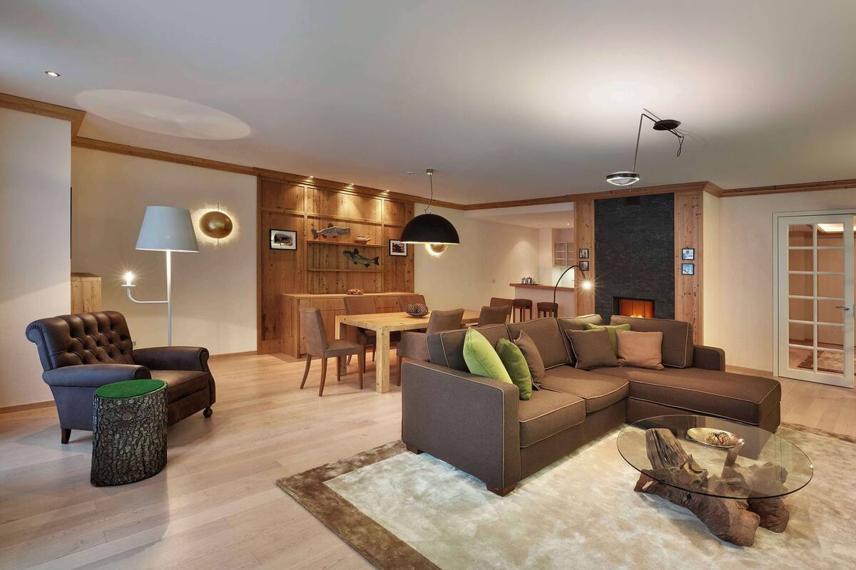 Una sala moderna con sofá, sillón y mesa de centro. Hay un comedor con mesa y sillas, y una zona de cocina con estufa. El espacio tiene pisos de madera y una chimenea.