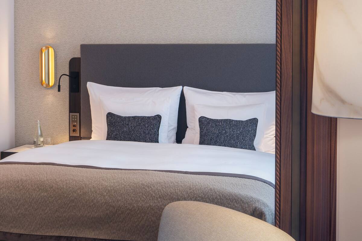 Una habitación de hotel con una cama grande, una silla, una lámpara de pared y una pared con textura.