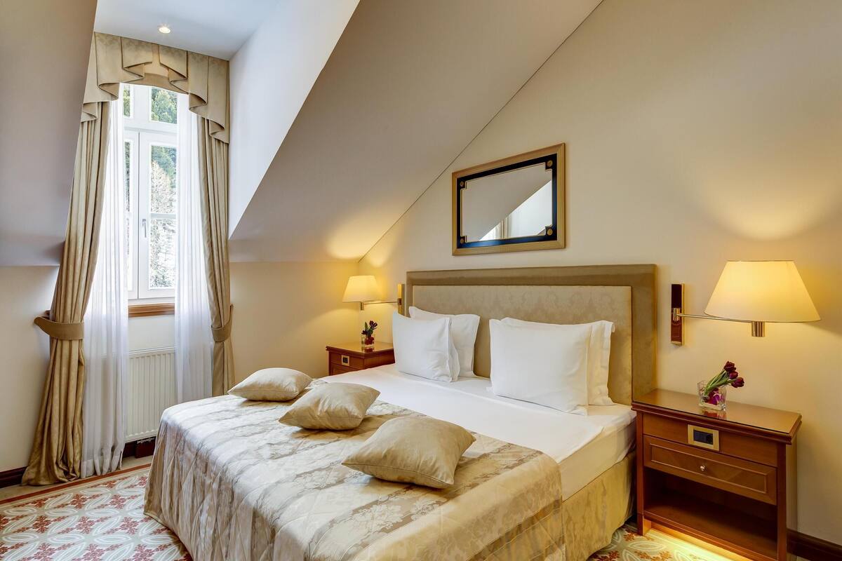 Un dormitorio ordenado con una cama grande, dos mesitas de noche con lámparas, una ventana con cortinas y una lámpara en el techo.