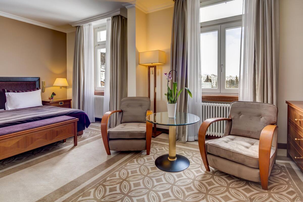 Habitación de hotel con cama, dos sillones, una mesita, lámpara y vistas a la ciudad a través de amplias ventanas.