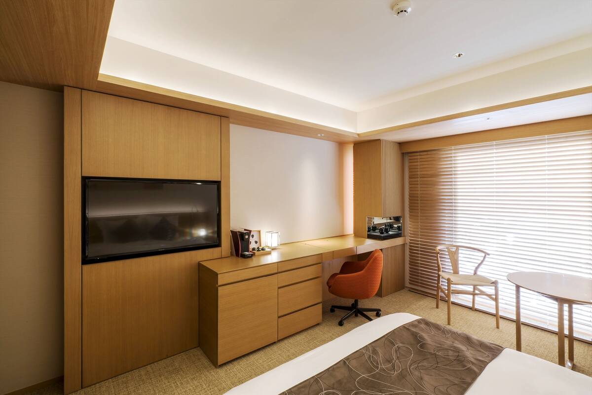 A modern hotel room with a wooden desk, a flat-screen TV, a comfortable orange chair, a small table, and a large window with blinds.