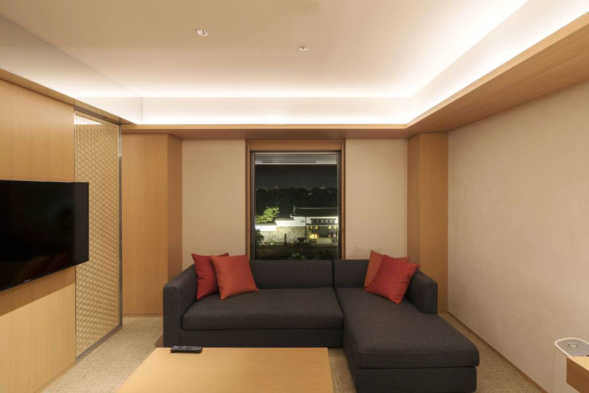 A modern living room with a sectional sofa, a flat-screen TV, and a window with a view of buildings.