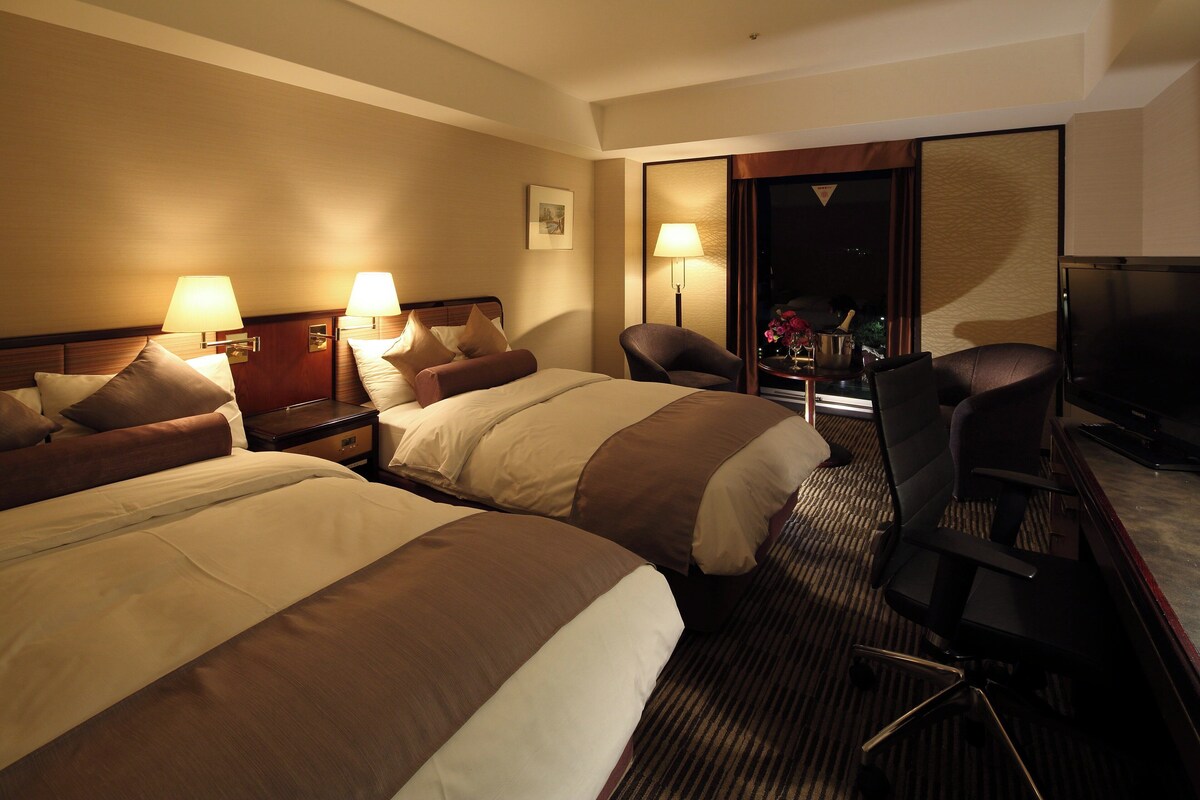 Une chambre d’hôtel avec deux lits, un bureau, une chaise, une télévision et une fenêtre.