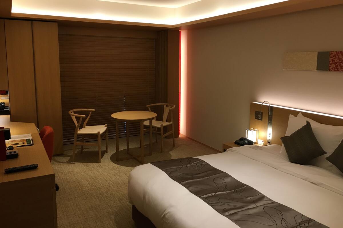 A modern hotel room with a bed, bedside table, lamp, and a seating area with a table and chairs.