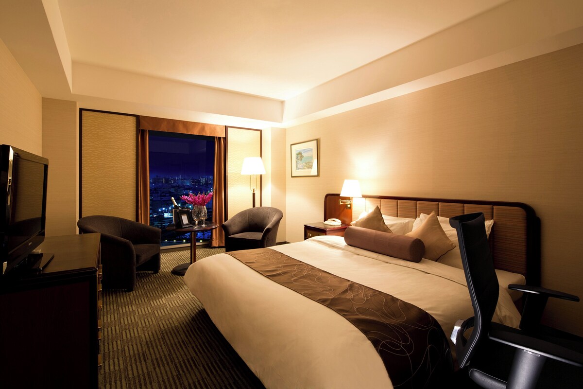 Une chambre d’hôtel équipée d’un lit, d’un bureau, d’une chaise, d’une télévision, d’une table de chevet et d’une fenêtre donnant sur la ville.