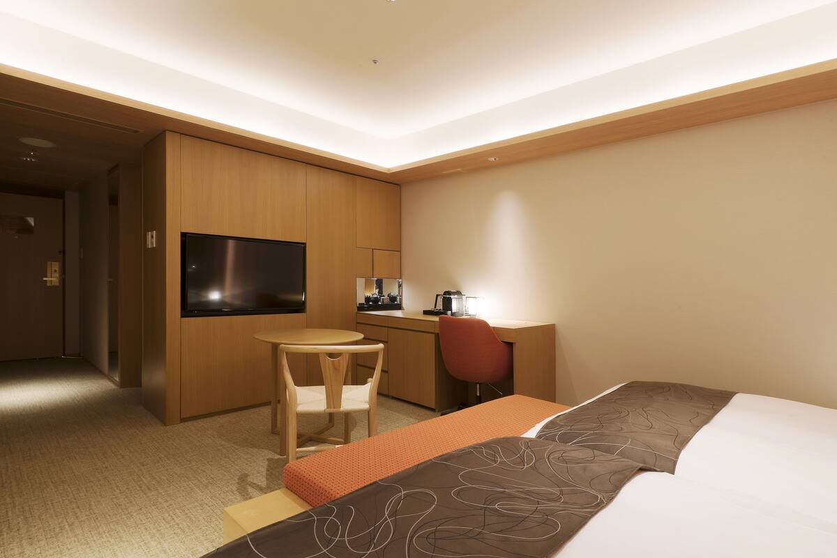 A hotel room with a bed, a desk, a chair, a television, and a kitchenette.