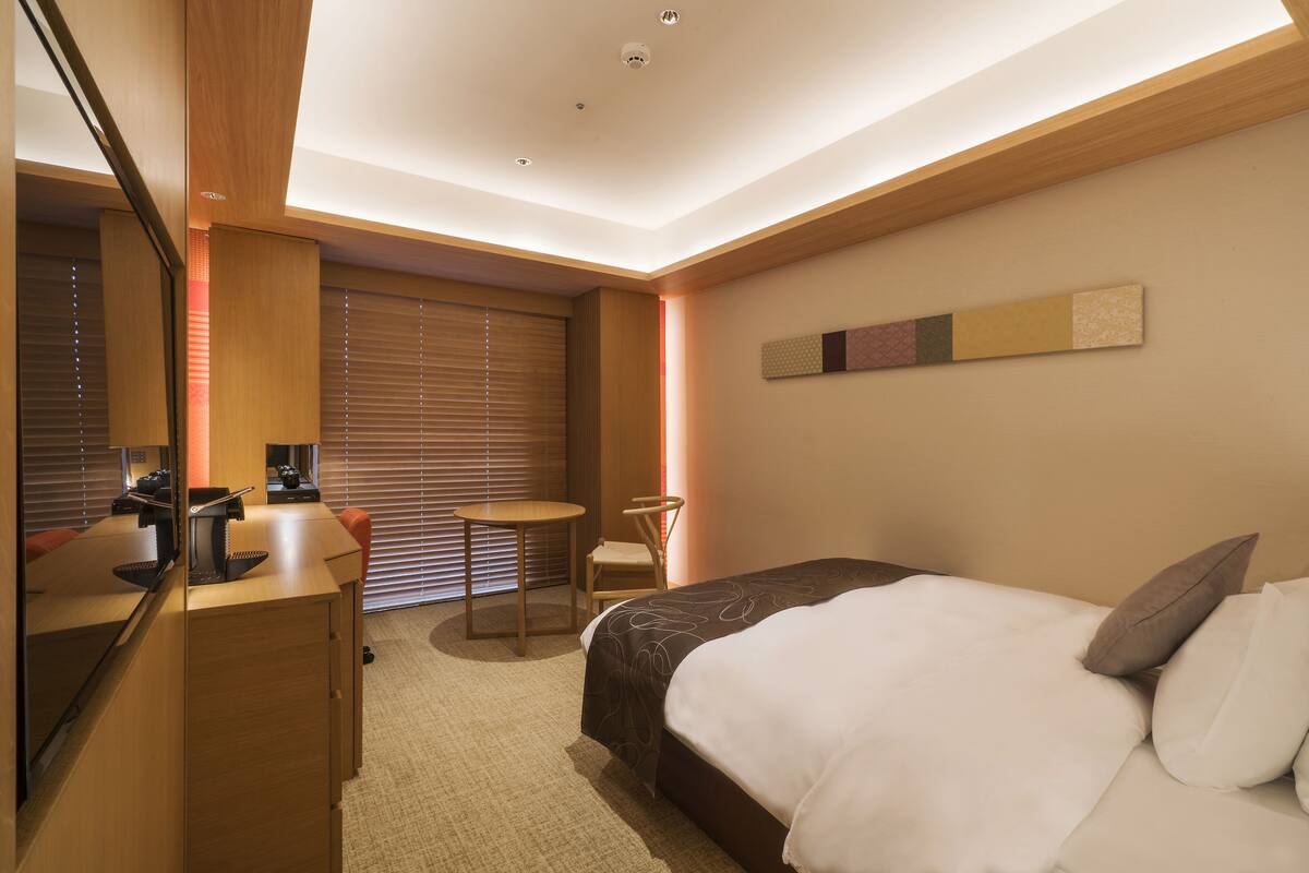 A hotel room with a bed, a desk, a chair, and a large window with blinds.
