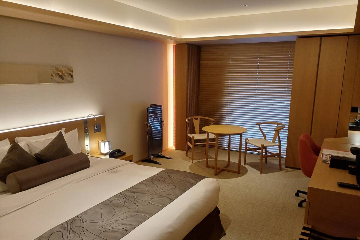 A hotel room with a bed, bedside table, lamp, chair, table, and a desk with a chair.