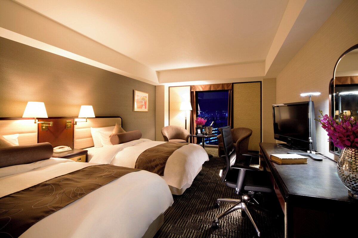 Une chambre d’hôtel avec deux lits, un bureau avec un ordinateur, une télévision et une vue sur la ville la nuit.