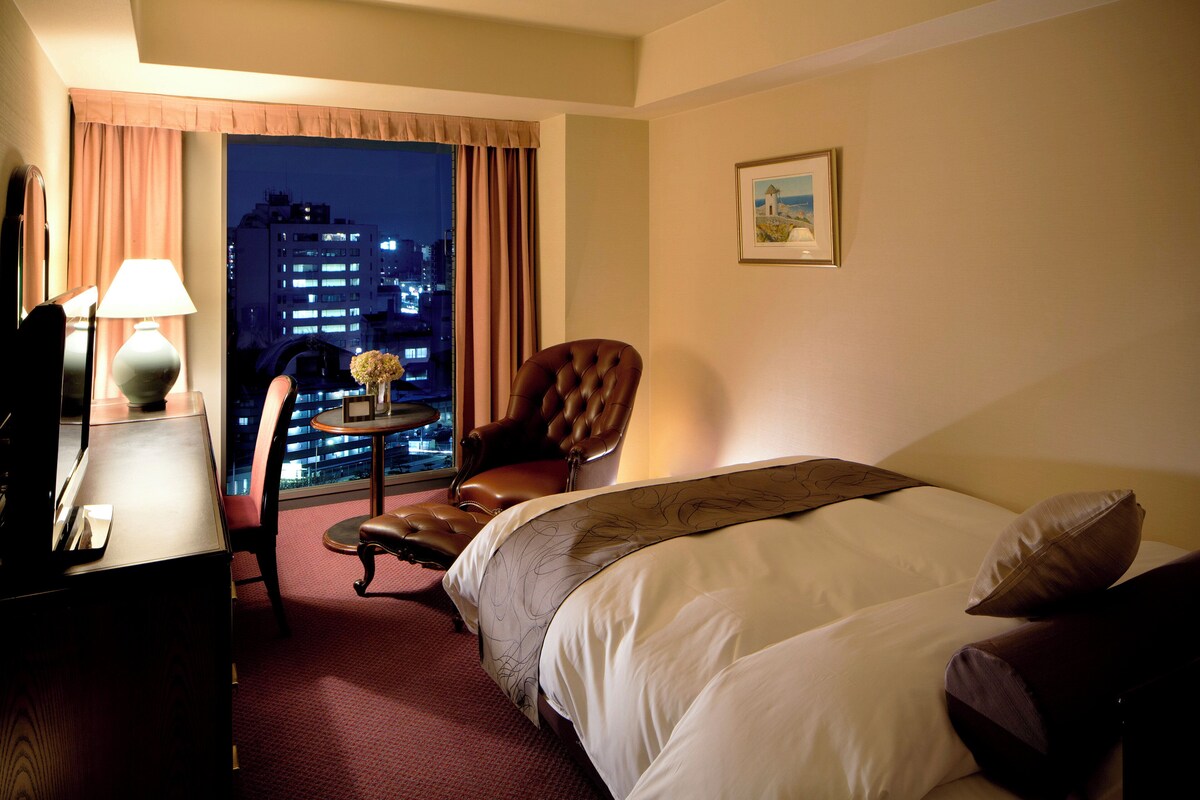Une chambre d’hôtel avec un lit, une chaise, un bureau avec une lampe, une petite table avec des fleurs et une vue sur la ville par la fenêtre.