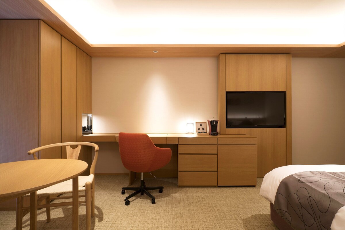 Une chambre d’hôtel avec un bureau en bois, une chaise de bureau rouge, un lit recouvert d’une couvre-lit à motifs et un téléviseur fixé au mur.